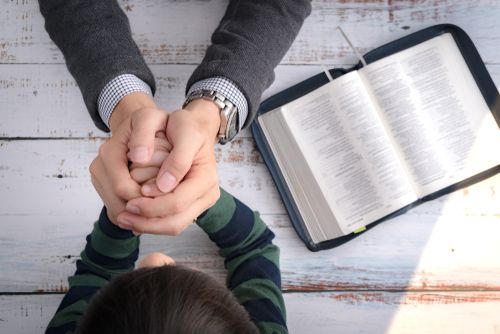 An adult clasps hands with a child while a religious book sits open on a table next to them