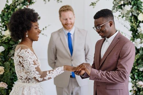 A groom places a ring on the bride's finger as the wedding officiant looks on.